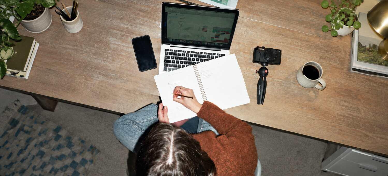 A person writing in a notebook beside their laptop